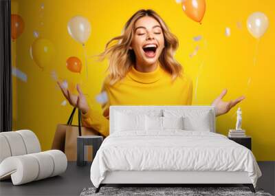 Portrait of wow excited amazed woman with open mouth holding paper shopping bag with colored helium balloons on a yellow background Wall mural