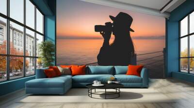 Silhouette of a woman photographer in hat with dslr camera during taking pictures of sea at sunset Wall mural