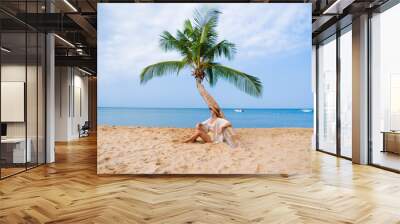 Young calm woman traveler sitting under a palm tree while relaxing on a tropical sandy island and enjoying idyllic and beautiful moment life Wall mural