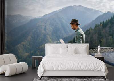 young man freelancer traveler wearing hat anywhere working online using laptop and enjoying mountain Wall mural