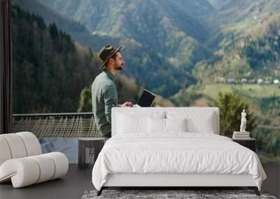 Young man freelancer traveler wearing hat anywhere working online using laptop and enjoying mountains view Wall mural