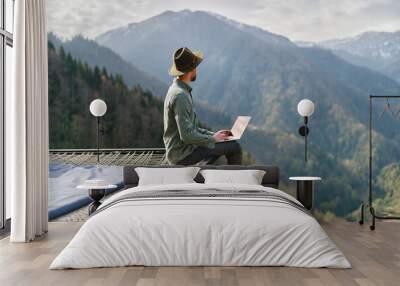 Young man freelancer traveler wearing hat anywhere working online using laptop and enjoying mountains view Wall mural