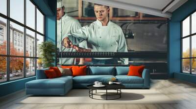 Food concept. Young handsome chef in white uniform monitors the degree of roasting and turns meat with the forceps in interior of restaurant kitchen. Preparing traditional beef steak on barbecue oven. Wall mural