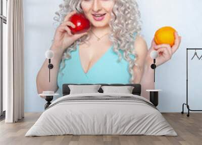 cute young girl with curly white hair and plump figure holding apple and orange fruit on studio background Wall mural