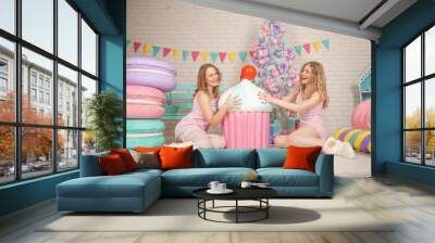 two cute best friends sit in pink pajamas on the floor, enjoy and laughing near the Christmas tree with gifts Wall mural