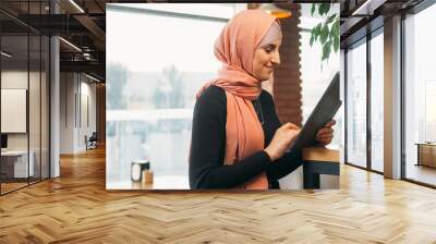 Cafe freelance. Internet user. Cheerful woman in hijab chatting on digital tablet web browsing social network at cozy coffee shop copy space. Wall mural