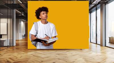 Doubtful black teenager. Think idea. Puzzled african male student writing in notebook isolated on orange copy space. Educational task. Find decision. Knowledge and skills Wall mural