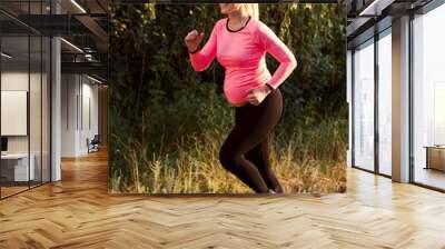 Happy pregnant woman jogging outside. Smiling expectant blonde running in forest. Sport, pregnancy, healthy lifestyle concept Wall mural