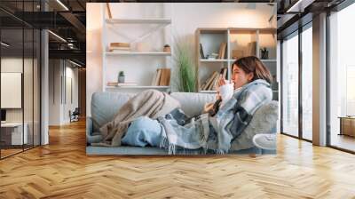 Sick tea. Virus drinking. Sore throat. Shivering woman catching cold suffering influenza holding cup healing grippe sitting wrapped blanket on couch in living room at home. Wall mural