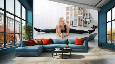 Stretching workout. Female aerobic fitness. Online training. Home sport. Happy slim woman in black sportswear doing bending forward legs apart looking laptop in light room interior. Wall mural