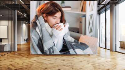 Tea warm. Rest drinking. Autumn shivering. Cold brunette woman in blanket enjoying hot beverage feeling frozen sitting on couch in light home interior. Wall mural