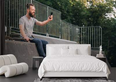 Urban man leisure. Youth lifestyle. Smiling bearded hipster sitting on ramp with skateboard and taking selfie. Wall mural