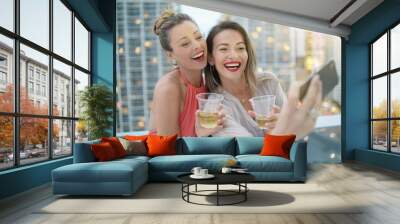 Two beautiful friends taking selfie on rooftop bar with city lights backdrop Wall mural