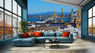 A view of Cádiz with industrial cranes, ships, and whitewashed rooftops, showcasing the city's coastal charm and maritime industry against a backdrop of mountains and blue sky. Wall mural