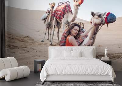 Gorgeous Caucasian woman wearing an orange long dress sitting on the sand with camels in a desert Wall mural