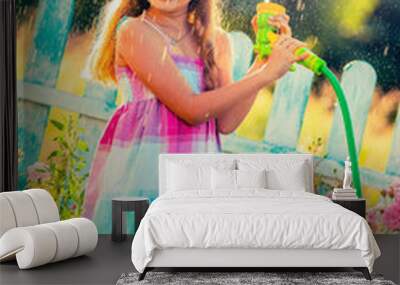 Playfull girl watering flowers with rain in the garden at summer day. Child using garden hose on sunny day at sunset lite. Little gardener playing in garden. Wall mural