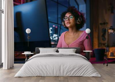 Attractive Multiethnic Latin Woman in Headphones Using Computer in Stylish Loft Apartment in the Evening. Creative Female Smiling, Browsing Videos on Social Media. Urban City View from Big Window. Wall mural