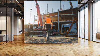 Back Shot of Worker Contractor Wearing Hard Hat and Safety Vests Walks on Industrial Building Construction Site. In the Background Crane, Skyscraper Concrete Formwork Frames  Wall mural