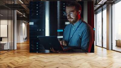 Bearded IT Specialist in Glasses is Working on Laptop in Data Center while Standing Near Server Rack. Running Diagnostics, Doing Maintenance Work. Emergency Red Light from Side Illuminating Specialist Wall mural