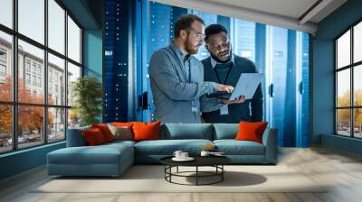 Bearded IT Technician in Glasses with Laptop Computer and Black Male Engineer Colleague are Using Laptop in Data Center while Working Next to Server Racks. Running Diagnostics or Doing Maintenance Wor Wall mural