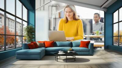 Beautiful and Creative Young Woman Sitting at Her Desk Using Laptop Computer. In the Background Bright Office where Diverse Team of Young Professionals Work. Wall mural