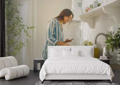 Beautiful Female is Using a Smartphone in a Kitchen While Preparing a Cup of Freshly Brewed Coffee from a French Press. Girl in Pyjamas with Healthy Creative Lifestyle Relaxes at Home in the Morning. Wall mural