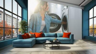 Beautiful Smiling Brunette Young Woman Sits in Front of a Washing Machine in Homely Jeans Clothes. She Loads the Washer with Dirty Laundry. Bright and Spacious Living Room with Modern Interior. Wall mural