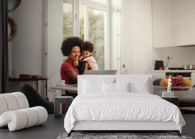 Beautiful Young Mom Holding Her Lovely Baby Son, Using Laptop Computer in Living Room in Apartment. Mother is Working from Home, Online Shopping, Watching Social Media or Writing Emails. Wall mural