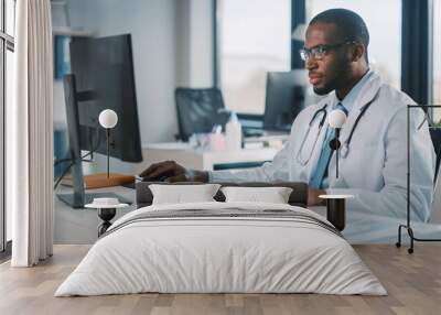 Calm African American Family Medical Doctor in Glasses is Working on a Desktop Computer in a Health Clinic. Physician in White Lab Coat is Browsing Medical History Behind a Desk in Hospital Office.  Wall mural