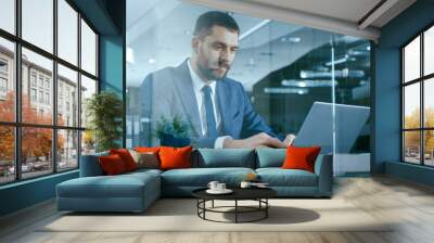 Confident Handsome Businessman Works on a Laptop at His Desk. Stylish Man in Modern Glass Office. Wall mural