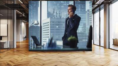 Confident Young Businessman in a Suit Standing in Modern Office, Talking on a Phone, Looking out of the Window on Big City with Skyscrapers. Successful Finance Manager Planning Work Projects. Wall mural