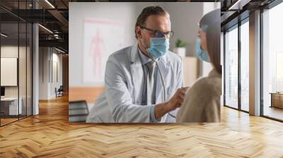 Doctor's Consultation Office: Physician Uses Stethoscope to Listen to Heartbeat and Lungs of the Female Patient. Medical Health Care Professionals Diagnosing, Treating Patient. Both Wearing Face Masks Wall mural