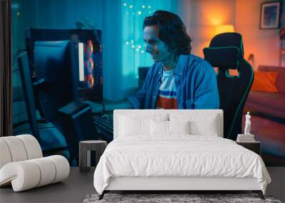 Excited Gamer Playing Online Video Game on His Personal Computer. Room and Personal Computer have Colorful Warm Neon Led Lights. Young Man has Long Hair and Handsome Smile. Cozy Evening at Home. Wall mural