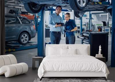 Female Mechanic Talking to a Manager Under a Vehicle in a Car Service. Specialist is Showing Info on a Tablet Computer. Empowering Woman Wearing Gloves and Safety Gloves. Modern Clean Workshop.  Wall mural