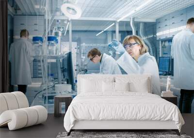 Female Scientist Analyzes Liquid in the Beaker and Types Down Observations on Her Computer. She's Working in a Busy Laboratory Full of Scientists Conducting Experiments. Wall mural