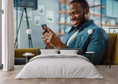 Handsome Black African American Man Using Smartphone while Sitting on a Sofa in Cozy Living Room. Freelancer Working From Home. Browsing Internet, Using Social Networks, Having Fun in Flat. Wall mural