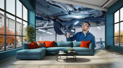 Handsome Professional Car Mechanic is Investigating Rust Under a Vehicle on a Lift in Service. Repairman is Using a LED lamp and Walks Towards. Specialist is Wearing Safety Glasses. Modern Workshop. Wall mural