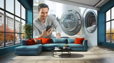 Handsome Smiling Young Man in Grey Jeans and Coat Sits in Front of a Washing Machine and Uses His Smartphone. He Loads Washer with Dirty Laundry. Bright and Spacious Living Room with Modern Interior. Wall mural