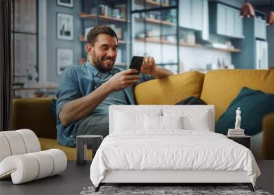 Happy Handsome Caucasian Man Using Smartphone in Cozy Living Room at Home. Man Resting on Comfortable Sofa. He's Browsing the Internet and Checking Videos on Social Networks and Having Fun. Wall mural