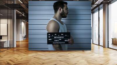 In a Police Station Arrested Beaten Man Poses for Side View Mugshot. He Wears Singlet, is Heavily Bruised and Holds Placard. Height Chart in the Background. Shot with Dark Cold Lights, Vignette Filter Wall mural