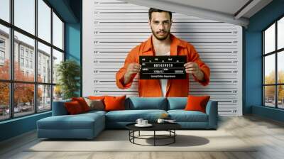 In a Police Station Arrested Man Getting Front-View Mug Shot. He's Wearing Prisoner Orange Jumpsuit and Holds Placard. Height Chart in the Background. Wall mural