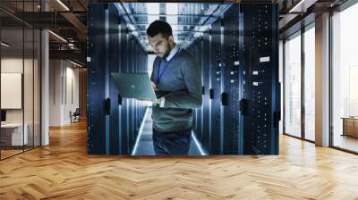 IT Technician Works on a Laptop in Big Data Center full of Rack Servers. He Runs Diagnostics and Maintenance, Sets up System. Wall mural