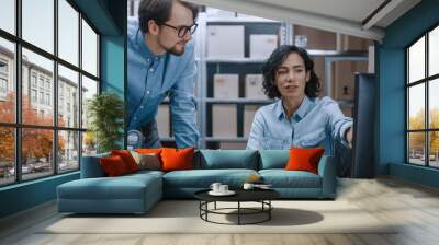 Male and Female Warehouse Inventory Managers Talking, Using Personal Computer and Checking Stock. Female Pointing at Screen. In the Background Rows of Shelves Full of Cardboard Box Packages. Wall mural