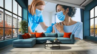 medical nurse in safety gloves and protective mask is making a vaccine injection to a female patient Wall mural