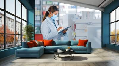 Pharmacy Drugstore: Portrait of Beautiful Pharmacist Wearing Face Mask Uses Digital Tablet Computer, Looks at Camera and Smiles Charmingly, Behind Her Shelves Full of Medicine Packages. Side View Shot Wall mural