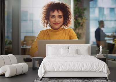 Portrait of a Beautiful Middle Eastern Manager Sitting at a Desk in Creative Office. Young Stylish Female with Curly Hair Looking at Camera with Big Smile. Colleagues Working in the Background. Wall mural