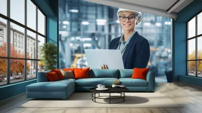 Portrait of a Happy Young Beautiful Female Engineer Wearing White Hard Hat, Using Laptop Computer in Office at Car Assembly Plant. Industrial Specialist Working on Vehicle Design in Modern Facility. Wall mural