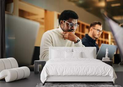 Portrait of Black Creative Young Man Arriving at Office for Work Office and Working on Laptop. Supervisor Smiling While Answering Email Questions From Colleagues. People Working in the Background Wall mural