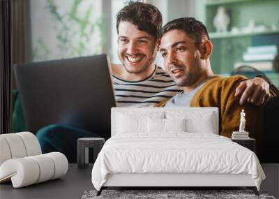 Portrait of Gentle Gay Couple Using Laptop Computer, while Sitting on a Couch in Cozy Stylish Apartment. Adult Boyfriends Online Shopping on Internet, Watching Funny Videos on Streaming Service. Wall mural