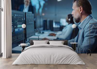 Portrait of Serious Professional Technical Controller Sitting at His Desk with Multiple Computer Displays Before Him. In the Background His Colleagues Working in System Control and Monitoring Center. Wall mural